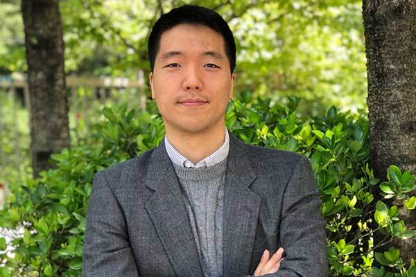 Korean male voice-over talent, wearing suit, poses outside studio with hedging in background.