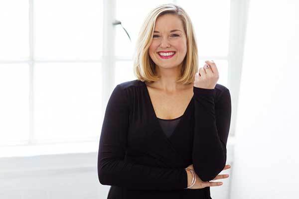 Blonde Irish female voice-over talent wearing all black, smiles broadly in a white, brightly lit room.