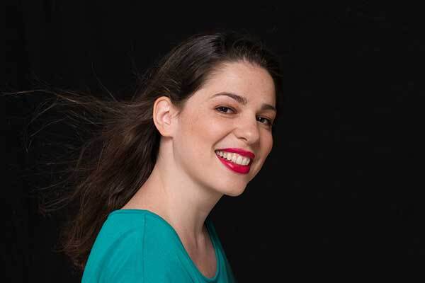 Dark-haired Greek female voice-over talent with red lipstick smiles at camera.